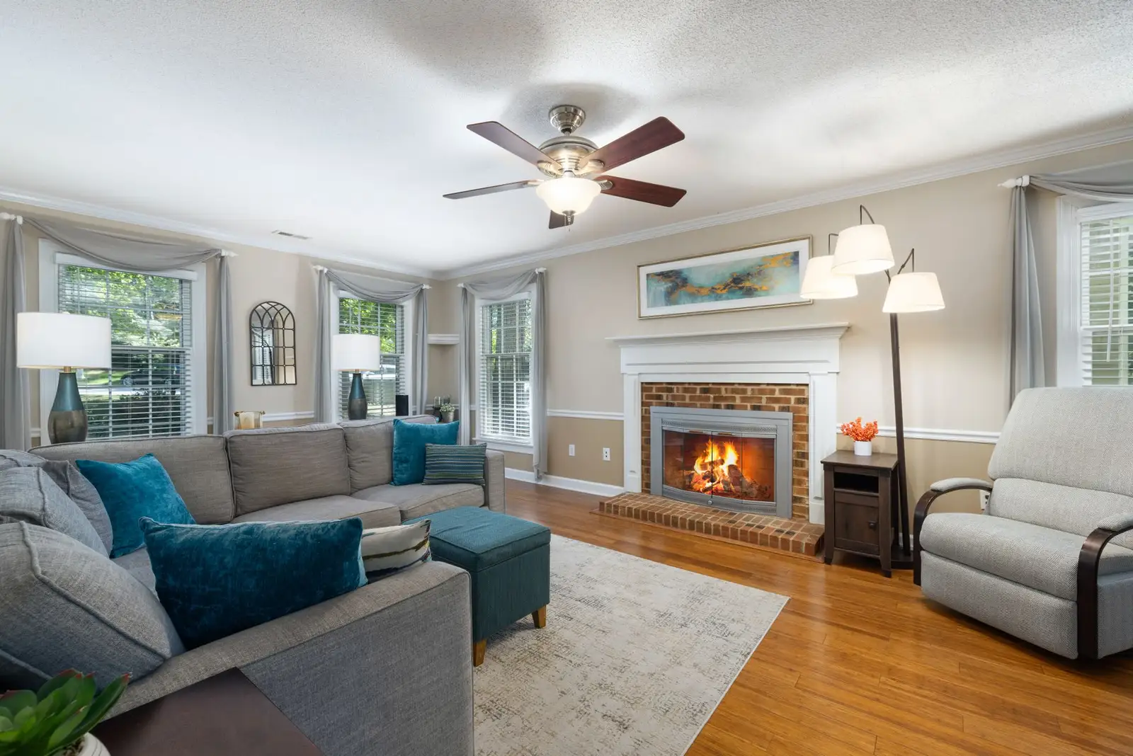 Living room with a grey sectional, teal cushions, brick fireplace and hardwood floors